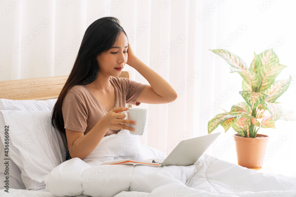 Freelance young woman using laptop working on internet holding cup of coffee in own hand while sitting on white bed with pillow in the morning at apartment. Lifestyle work at home, technology concept.
