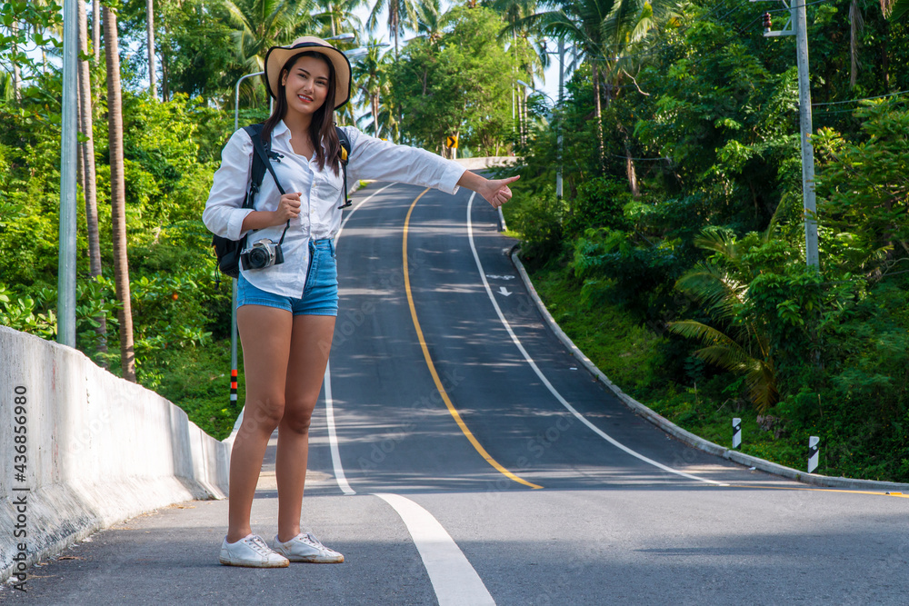 Adventure recreation and travel concept. Young woman wearing hat carrying backpack with camera use her hand hitchhiking for help while standing on the road.