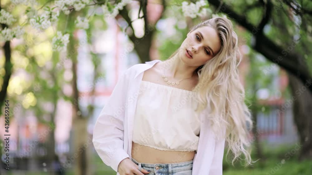 Attractive fashion model with flying hair in the flowered garden in the spring time. Young beautiful blonde woman shakes hair. Blossoms fall from trees. Slow motion Attractive fashion model with flying hair in the flowered garden in the spring time. Young beautiful blonde woman shakes hair. Blossoms fall from trees. Slow motion