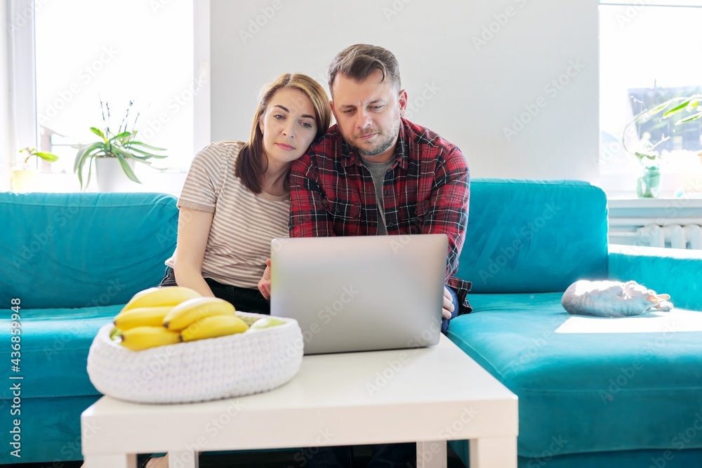 Serious middle-aged couple husband and wife with laptop sitting at home on couch