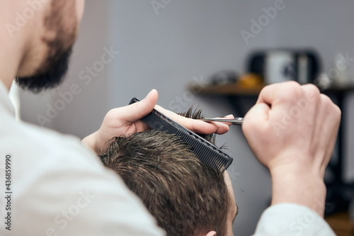 Professional barber making haircut to young man using scissors and comb at barbershop. Close-up, selective focus