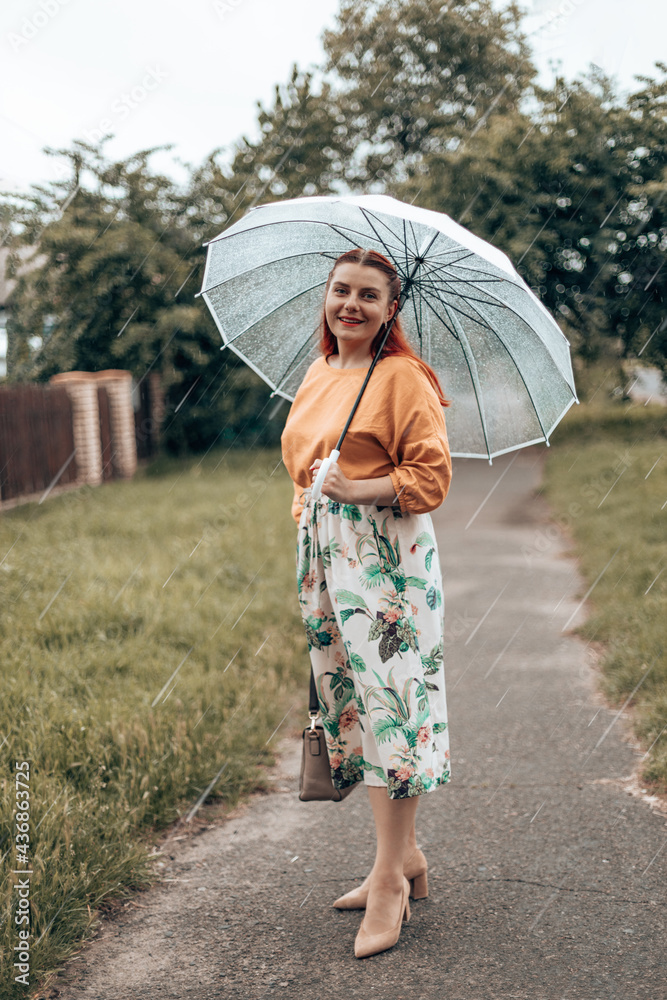 Happy young woman in bright clothes under a transparent umbrella in bad weather in the city park