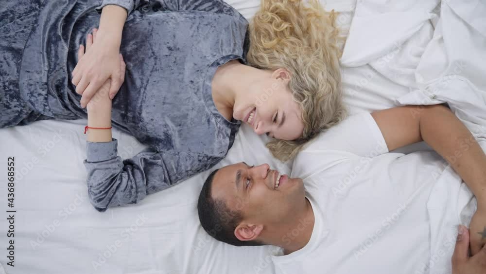 Top view of happy loving couple rubbing noses lying on bed in the morning laughing. Romantic African American man and Caucasian woman enjoying weekend together in bedroom Top view of happy loving couple rubbing noses lying on bed in the morning laughing. Romantic African American man and Caucasian woman enjoying weekend together in bedroom