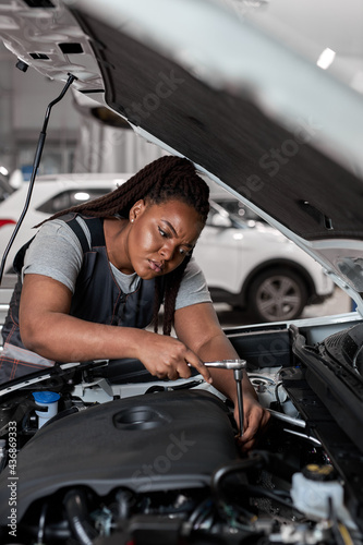 Beautiful Caucasian Female Mechanic is Working With Car In Car Service. Woman in Overalls is Working on Usual Car Maintenance, Using Ratchet. Modern Clean Workshop with Cars. © Roman