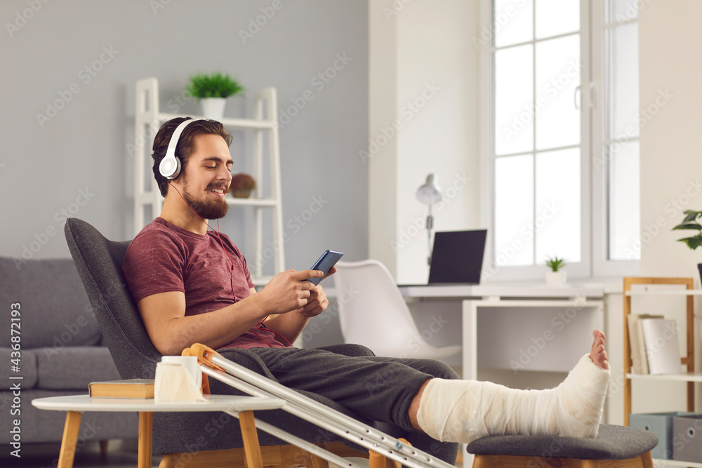Positive man enjoying sick leave despite physical injury. Happy smiling young guy with broken leg in plaster cast sitting in chair at home, using mobile phone and listening to music through headphones Positive man enjoying sick leave despite physical injury. Happy smiling young guy with broken leg in plaster cast sitting in chair at home, using mobile phone and listening to music through headphones