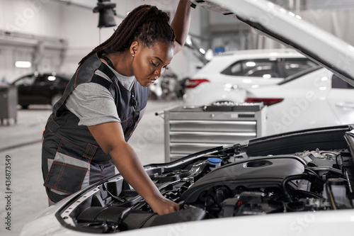 Afro Car mechanic woman is examining under hood of car at repair garage, wearing overalls, looking confident and concentrated. Side view on female trying to solve the problem of inoperative engine photo