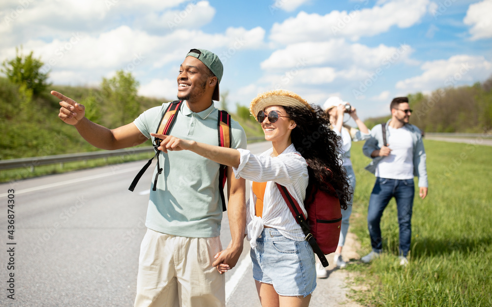 Millennial travelers with backpacks walking along road, hitchhiking in countryside. Group of people having autostop trip