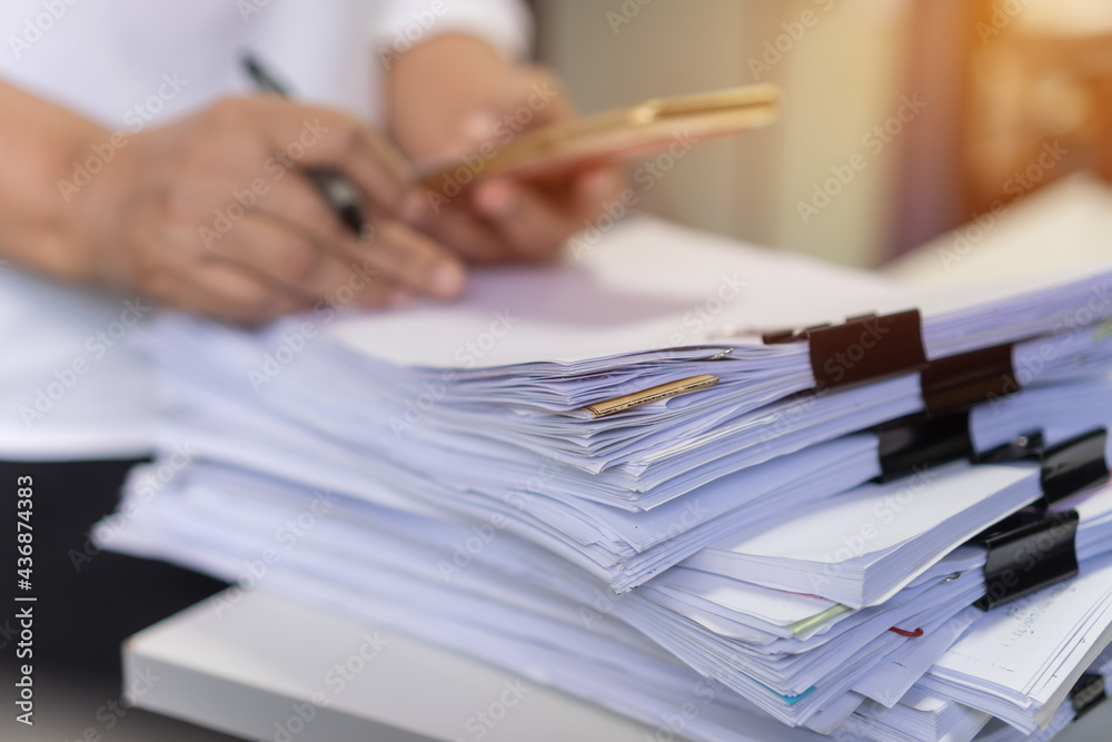 Paperwork files searching information business concept, Businessman hands using smartphone for working in stacks of report papers and piles of unfinished documents achieves on office desk Paperwork files searching information business concept, Businessman hands using smartphone for working in stacks of report papers and piles of unfinished documents achieves on office desk