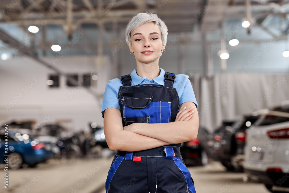 Caucasian professional female mechanic posing at camera standing in auto repair shop. Short haired woman in blue uniform. Car service, repair, maintenance and people concept. Front view. Copy space - obrazy, fototapety, plakaty 
