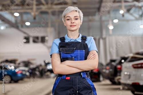 Caucasian professional female mechanic posing at camera standing in auto repair shop. Short haired woman in blue uniform. Car service, repair, maintenance and people concept. Front view. Copy space