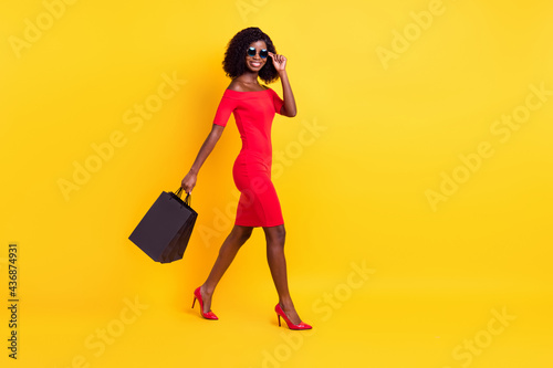 Full length photo of pretty shiny dark skin woman wear red clothes glasses walking holding bags isolated yellow color background
