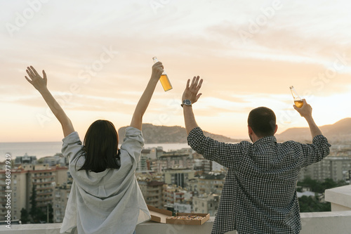 Cheerful couple enjoying the sunset seaside  drinking beer and eating pizza on the balcony. Happy people raise their hands up on the rooftop with cityscape back view.