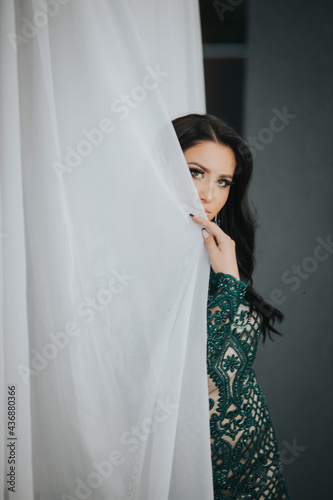 Young attractive lady in a fashionable green dress holding the curtain and posing photo