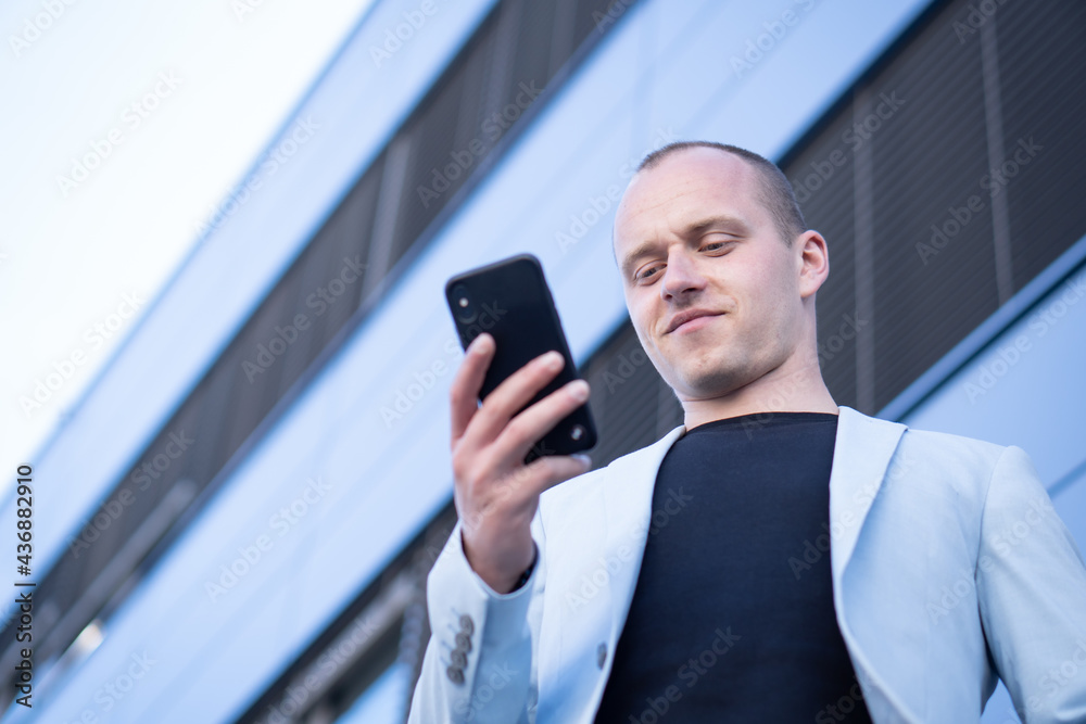 Bottom view perspective of young short-haired businessman in a blue suit standing in front of a corporate building and writes a text message on mobile. Bottom view perspective of young short-haired businessman in a blue suit standing in front of a corporate building and writes a text message on mobile.