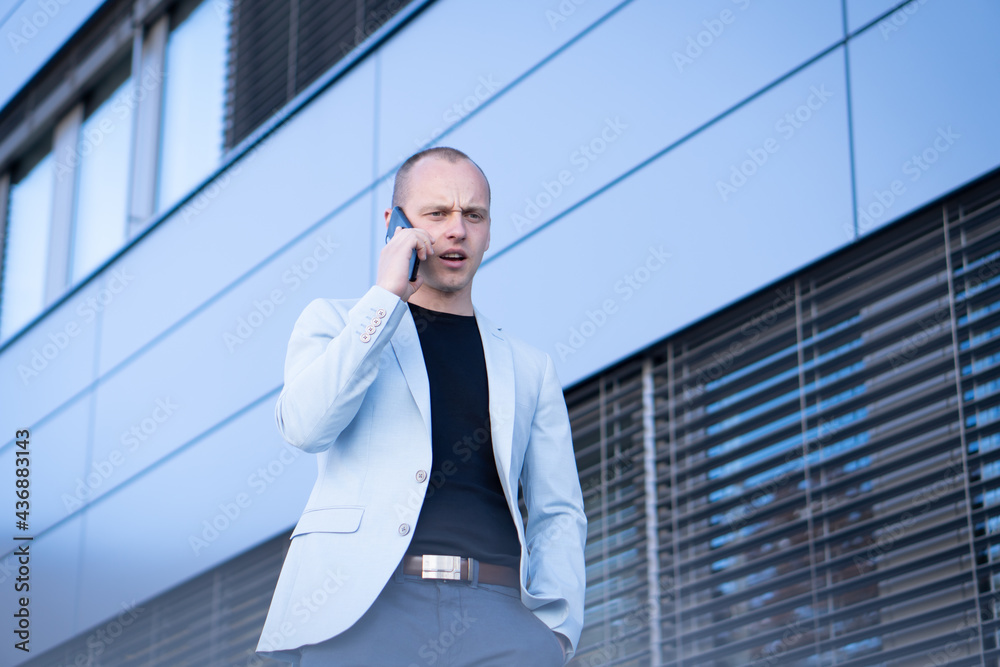 Bottom view perspective of young short-haired businessman in a blue suit standing in front of a corporate building solves a business phone call. Bottom view perspective of young short-haired businessman in a blue suit standing in front of a corporate building solves a business phone call.