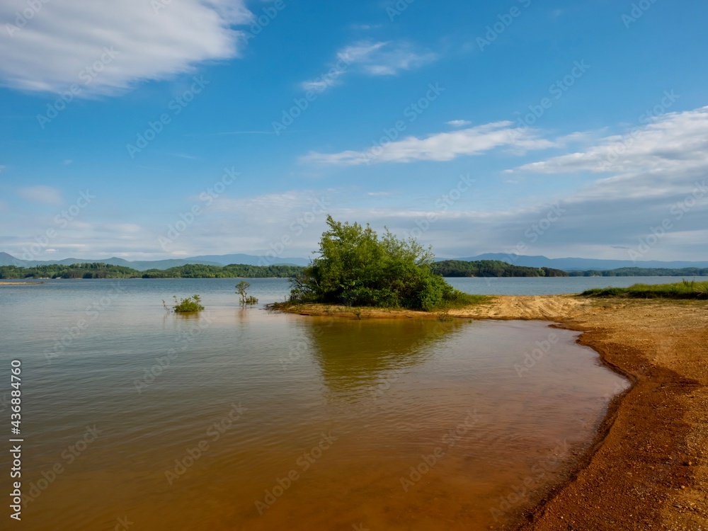 Douglas Lake in the spring in Tennessee with the Great Smoky Mountains in the background, the water and sandy shore with a blue sky filled with clouds in the distance.