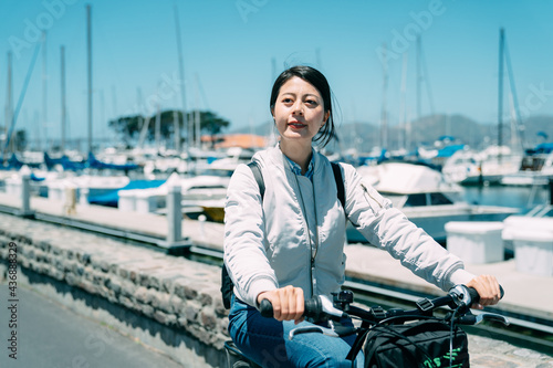 carefree asian woman is looking into the distance while enjoying riding a bike along marina with sailboats in Sausalito city, California on a sunny vacation day © PR Image Factory