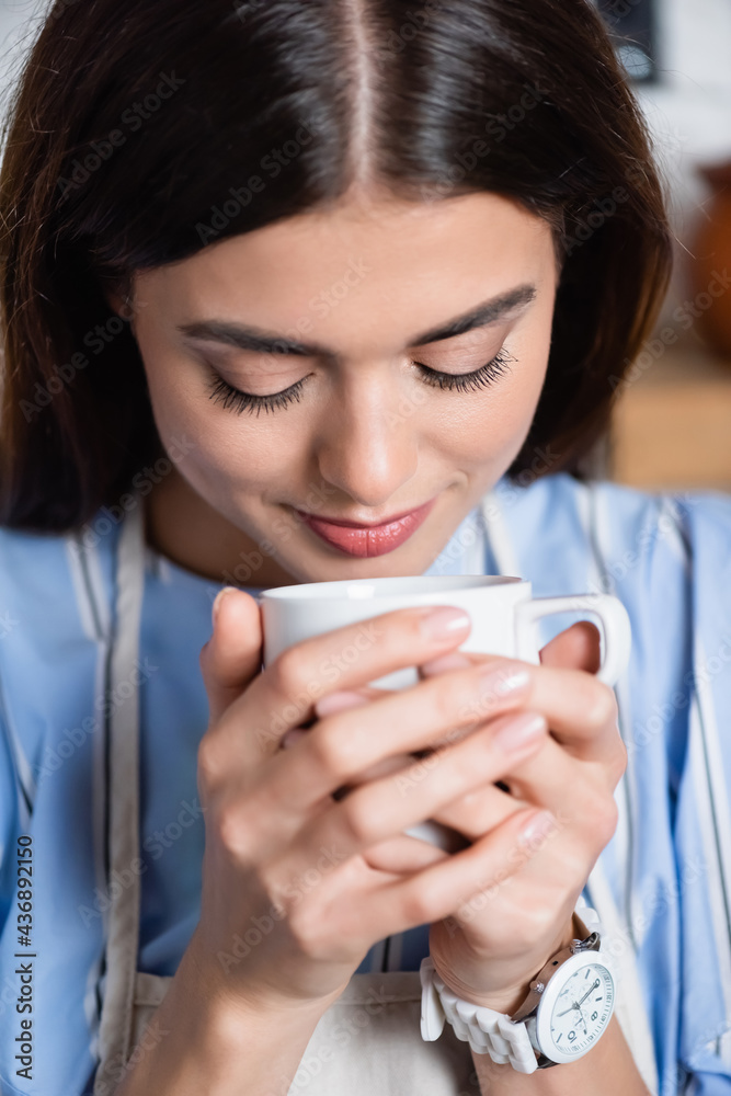 portrait of smiling young adult woman smelling aroma of coffee in white cup on blurred background portrait of smiling young adult woman smelling aroma of coffee in white cup on blurred background