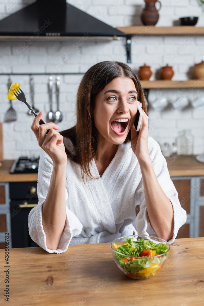 young adult woman in bathrobe eating vegetables salad and speaking on cellphone in modern kitchen