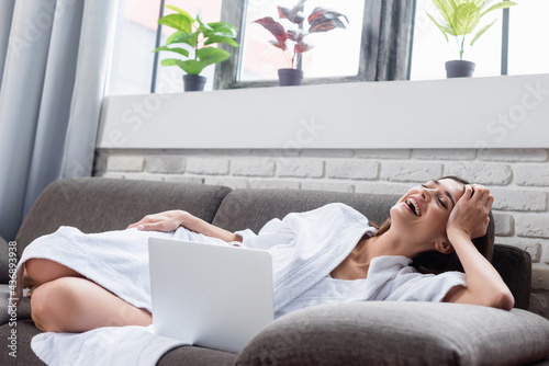 laughing young adult woman with closed eyes resting on couch near laptop at home