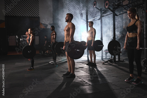Group of sporty muscular people working out in gym. Cross fit training. Handsome men and attractive women doing exercises with barbells. Weightlifting. In dark smoky space