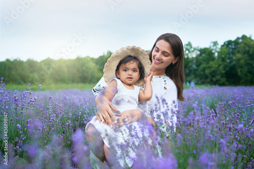 Young happy mother sitting between purple flowers with kid wearing straw hat and looking at camera. Smiling woman posing with baby daughter on knees in lavender field. Concept of nature, motherhood.