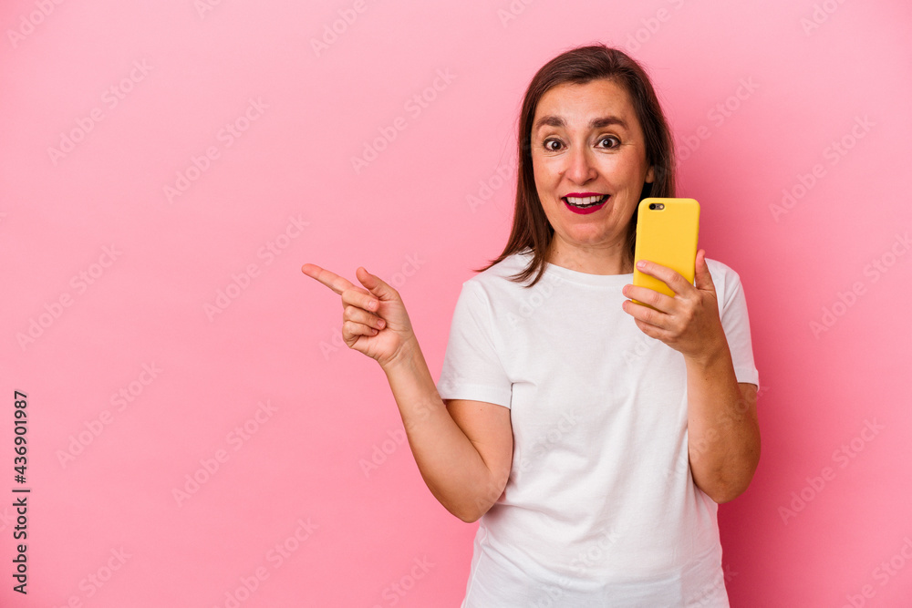 Middle age caucasian woman holding mobile phone isolated on pink background smiling and pointing aside, showing something at blank space. Middle age caucasian woman holding mobile phone isolated on pink background smiling and pointing aside, showing something at blank space.