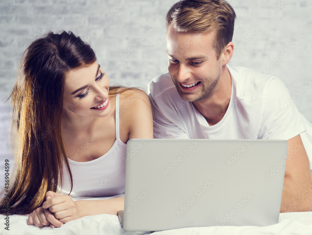Beautiful young happy smiling couple using laptop, on bed, against white bricks loft style wall. Internet, technology, family, love, relationship concept.