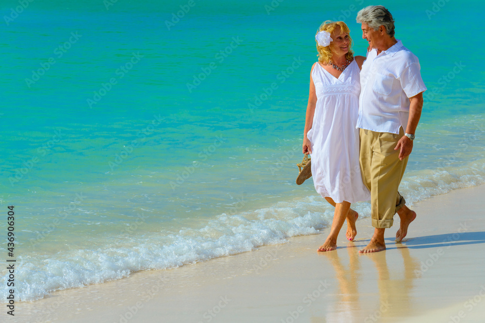 Older couple hugging and walking on the beach in white outfits Older couple hugging and walking on the beach in white outfits
