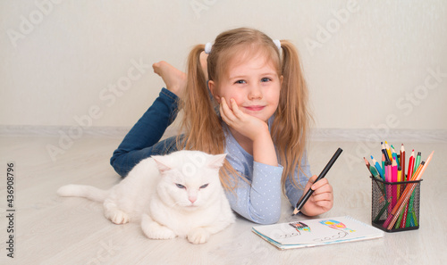 Little smilling playfull girl lying on the floor with her white cat and drawing a house.