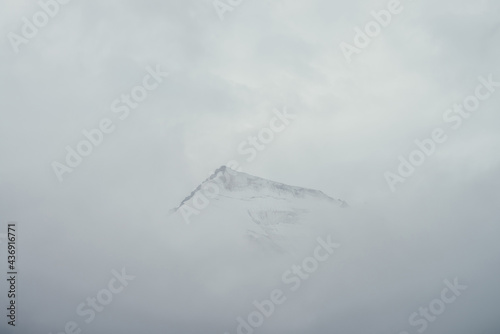 Minimal mountain landscape with high pointy rock in clouds. Minimalist mountain scenery with sharp snowy mountain peak over clouds. Snow-white pointed pinnacle above white clouds. Big top in dense fog