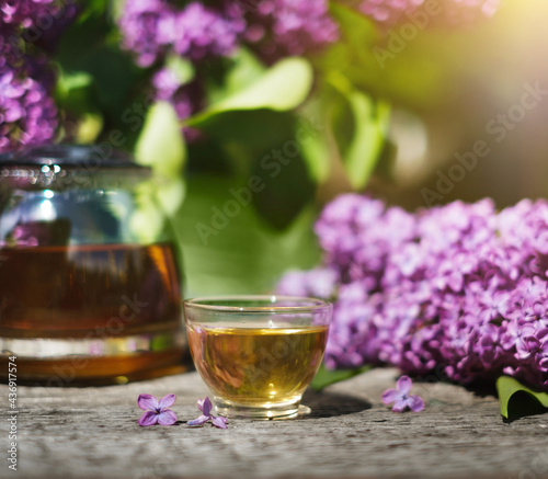 A glass teapot with herbal tea in the background and a glass bowl on a wooden table against a background of lilac flowers on a sunny summer day. The concept of medicinal herbal tea for health.