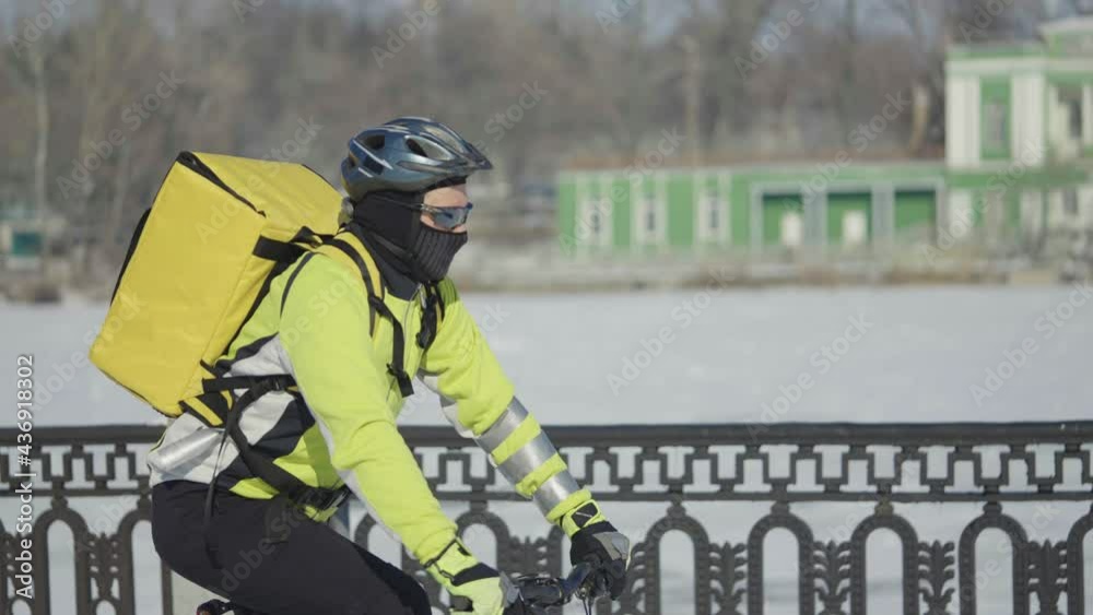 Man with large bag in helmet rides bicycle along snowy street and delivers food or parcels. Fast courier delivery in city. Extreme working conditions on slippery winter road. Slow motion. Close up. Man with large bag in helmet rides bicycle along snowy street and delivers food or parcels. Fast courier delivery in city. Extreme working conditions on slippery winter road. Slow motion. Close up.