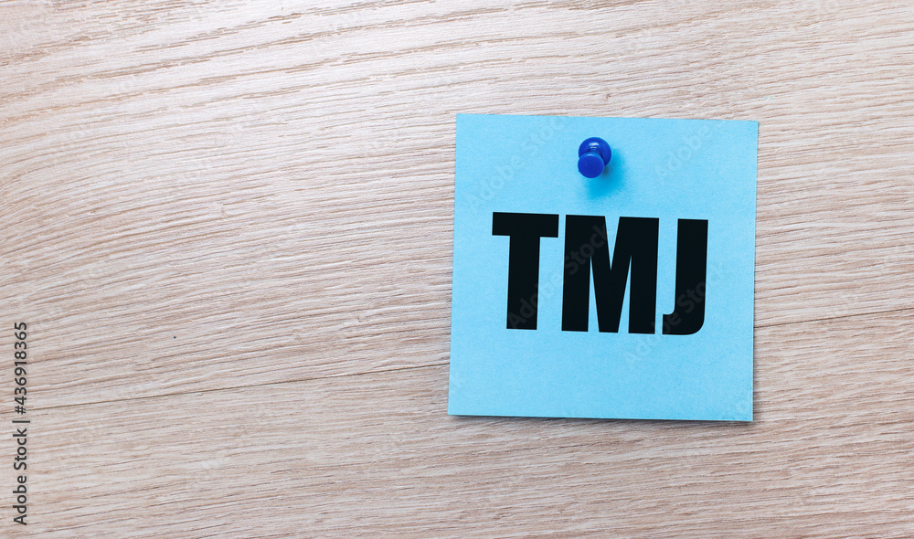 On a light wooden background - a light blue square sticker with the text TMJ On a light wooden background - a light blue square sticker with the text TMJ