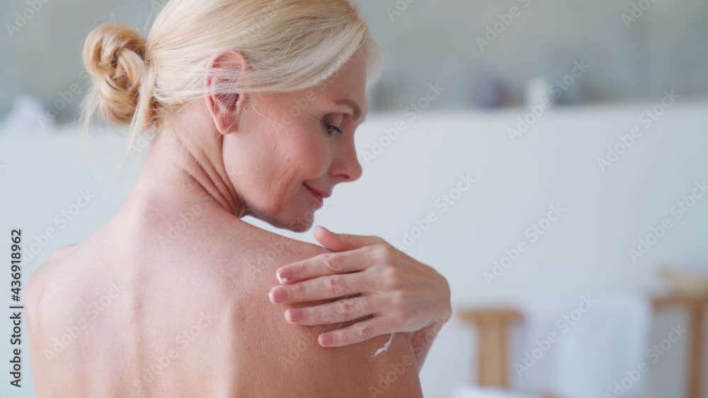 Back view of nude beautiful middle aged 50s woman applying moisturizing body lotion after shower. Advertising of bodycare spa procedures antiage delicate dry skin care products concept.