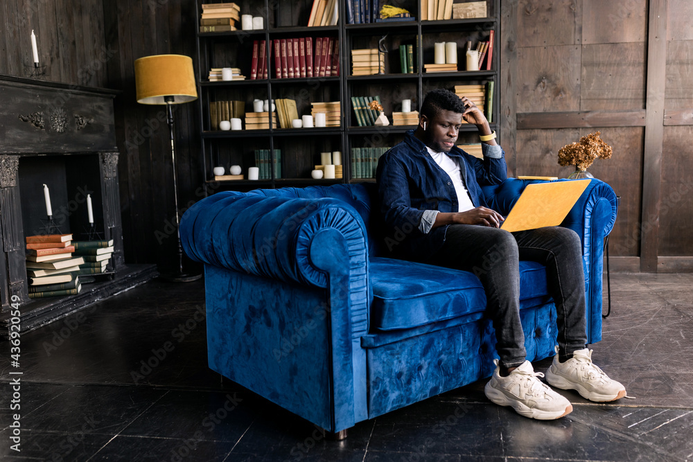 Naklejka premium Serious African american manager wear headphones looking at laptop screen provide support to client distantly, hold online conference. Student study distantly at modern blue and yellow apartment