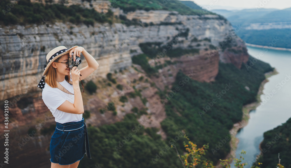 young blonde girl in summer hat takes photo on retro camera of panorama horizin mountain landscape walking  outdoors, hipster tourist enjoys hobby of photographing summer nature on holiday