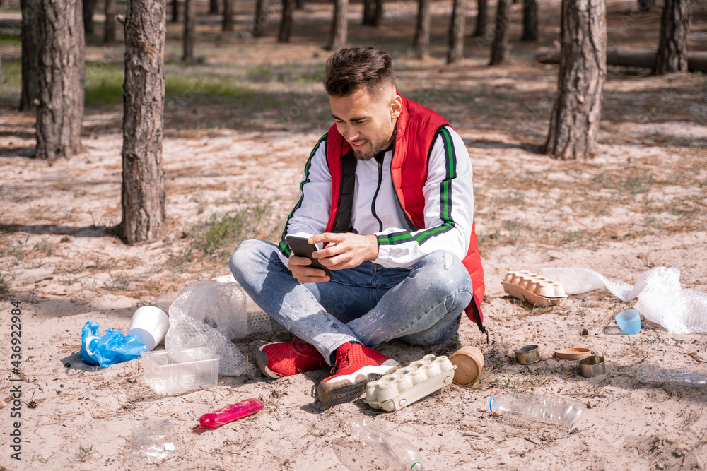 cheerful man with crossed legs sitting and using smartphone near trash on ground cheerful man with crossed legs sitting and using smartphone near trash on ground