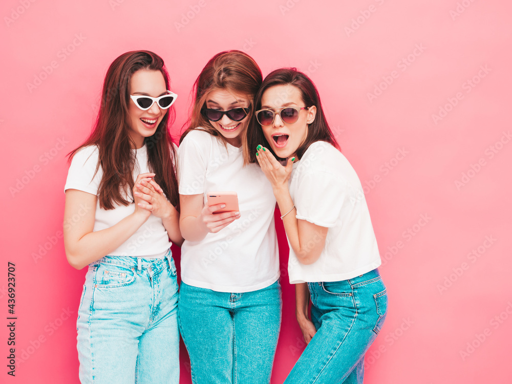 Three young beautiful smiling hipster female in trendy summer t-shirt and jeans clothes. Sexy carefree women posing near pink wall in studio.Positive models looking at smartphone screen. Using apps Three young beautiful smiling hipster female in trendy summer t-shirt and jeans clothes. Sexy carefree women posing near pink wall in studio.Positive models looking at smartphone screen. Using apps