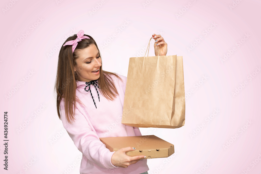 Beautiful young woman in a pink sweater (hoodie) holds a craft package and a box of pizza isolated on background. Beautiful young woman in a pink sweater (hoodie) holds a craft package and a box of pizza isolated on background.