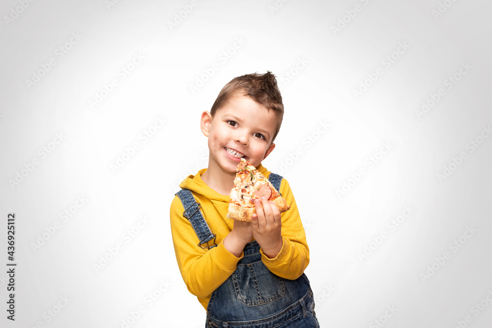 Little cute toddler boy in a yellow sweater and denim overalls holds a slice of pizza near his mouth and smiles beautifully. Little cute toddler boy in a yellow sweater and denim overalls holds a slice of pizza near his mouth and smiles beautifully.