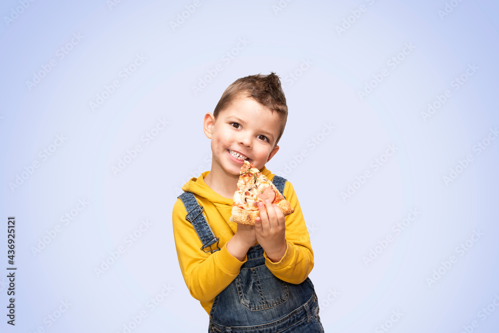 Little cute toddler boy in a yellow sweater and denim overalls holds a slice of pizza near his mouth and smiles beautifully. Little cute toddler boy in a yellow sweater and denim overalls holds a slice of pizza near his mouth and smiles beautifully.