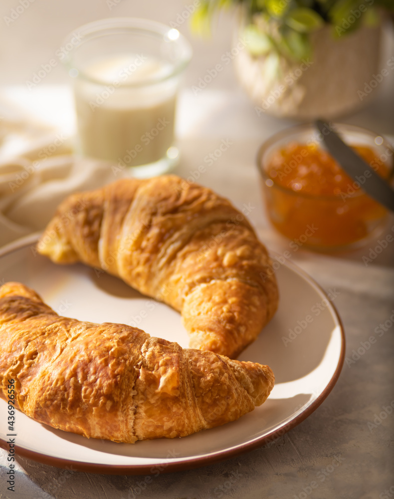 In the photo we see a still life of two ruddy croissants on a white plate, golden orange jam and a glass of milk. Pastel shades. White background. Bright lighting. There are no people in the photo.