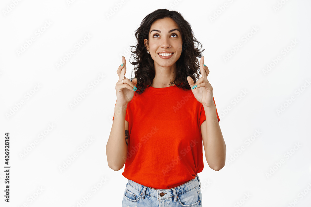 Image of young woman praying, looking up with fingers crossed, making wish, hope for good luck, standing in red t-shirt against white background Image of young woman praying, looking up with fingers crossed, making wish, hope for good luck, standing in red t-shirt against white background