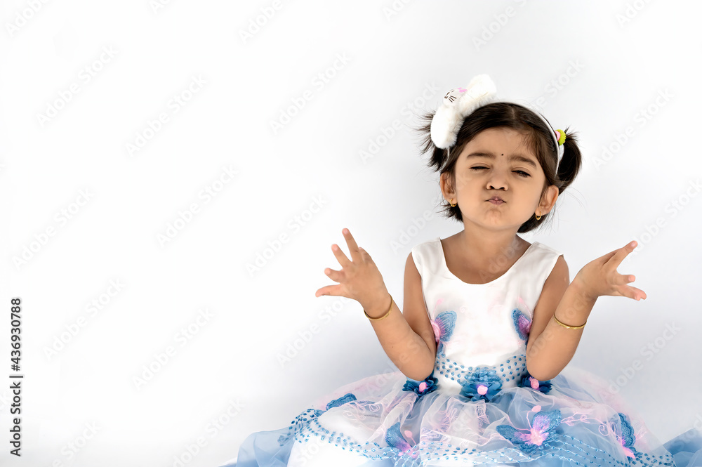Cheerful Indian / Asian kid with shrug gesture asking something and looking at camera. Portrait of little girl in denim overalls. Isolated on white. Question concept.