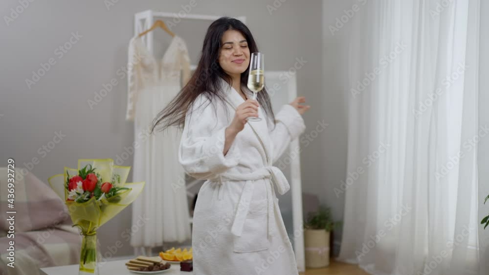 Joyful modern Middle Eastern bride dancing spinning with champagne glass drinking alcohol on wedding morning in living room. Portrait of happy excited young woman enjoying bridal preparations