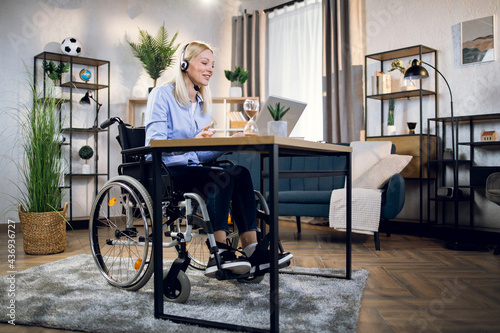 Pretty young woman in wheelchair using headset and laptop for video conference at home. Handicapped business lady leading working meeting online.