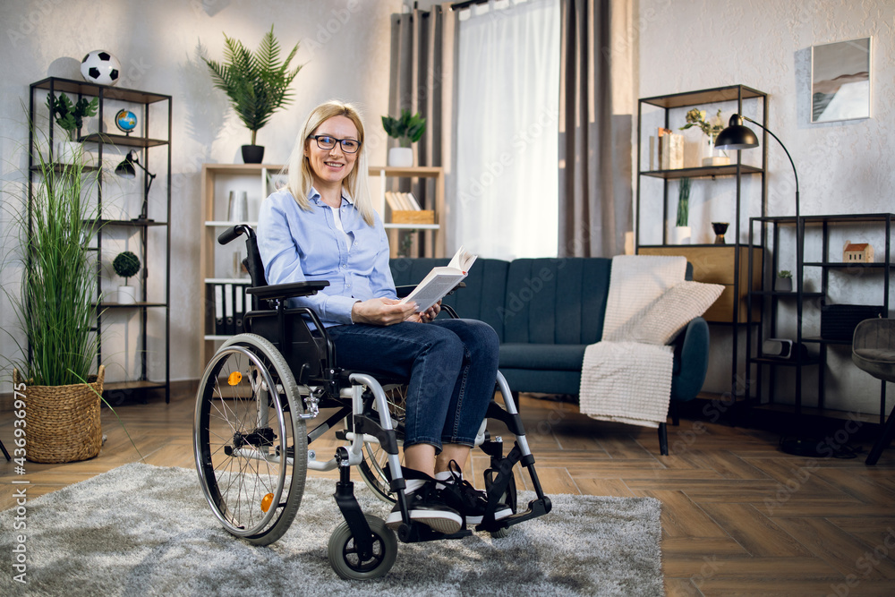 Positive young woman in eyewear and casual outfit smiling on camera while sitting in wheelchair with book in hands. Concept of people and literature. Positive young woman in eyewear and casual outfit smiling on camera while sitting in wheelchair with book in hands. Concept of people and literature.
