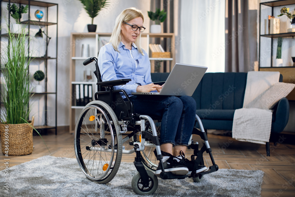 Pleasant young woman with disabilities working on modern laptop while staying at home. Charming blonde in eyewear and casual clothes typing on keyboard while sitting in wheelchair. Pleasant young woman with disabilities working on modern laptop while staying at home. Charming blonde in eyewear and casual clothes typing on keyboard while sitting in wheelchair.
