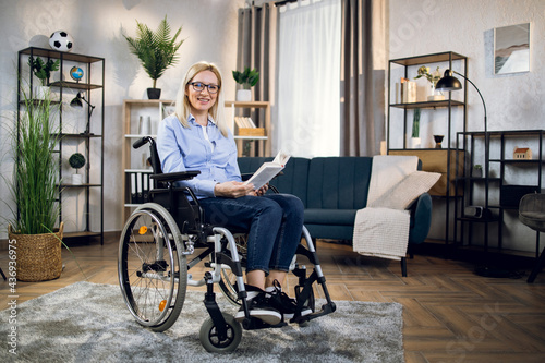 Positive young woman in eyewear and casual outfit smiling on camera while sitting in wheelchair with book in hands. Concept of people and literature.